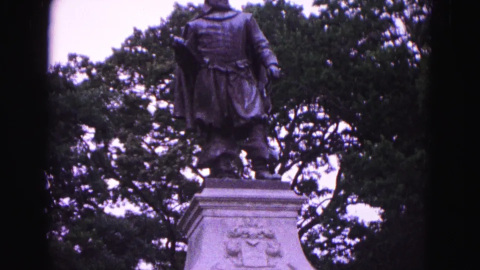 1964 Williamsburg home-movie frame showing the Captain John Smith statue above a pale stone pedestal in a tree-filled park.