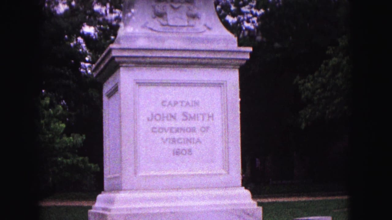 The monument pedestal fills the frame, with the Captain John Smith inscription visible against trees and lawn in Williamsburg.