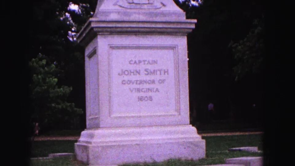 A closer handheld view of the Captain John Smith pedestal shows the carved inscription, stone base, trees, and lawn.