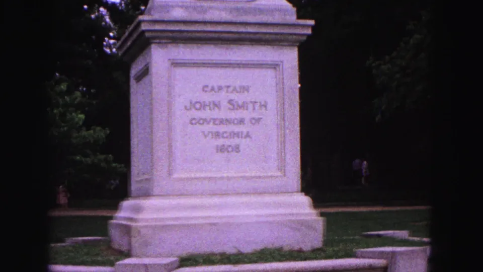 1964 Williamsburg home-movie frame showing the Captain John Smith monument pedestal with inscription, grass, trees, and distant visitors.