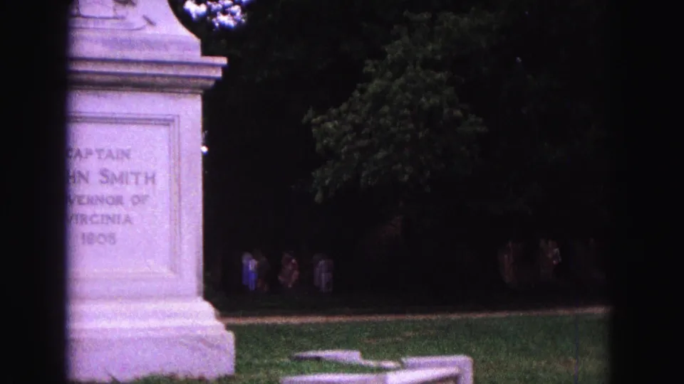 Late frame shows the Captain John Smith pedestal partly cropped, with the park lawn, mature trees, and distant visitors still visible.