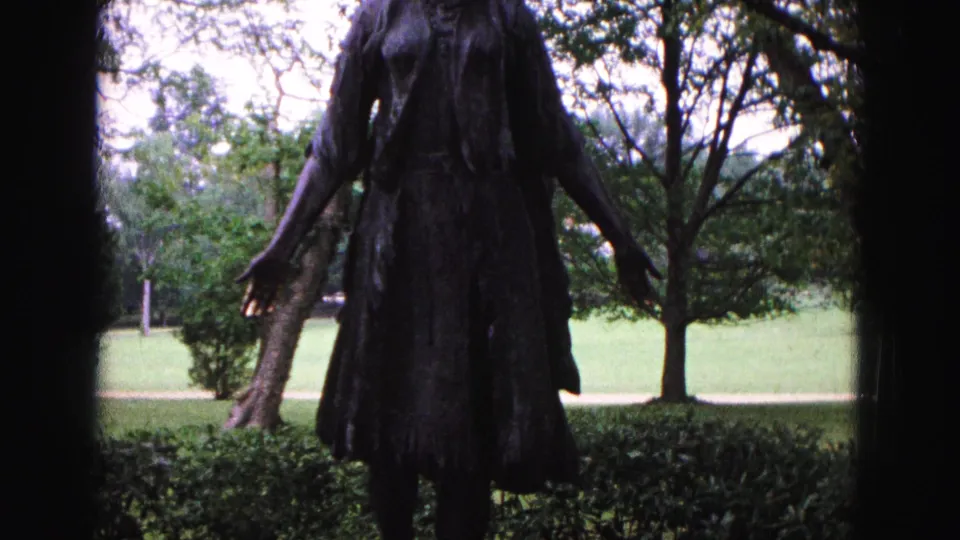 1964 Williamsburg home-movie frame with the bronze woman statue centered from torso toward skirt against trees and grass.