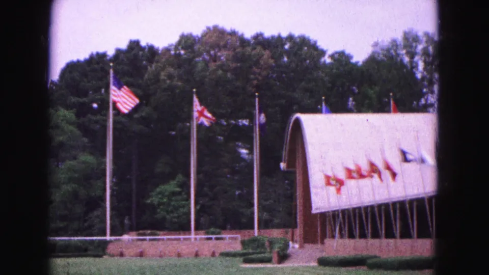 1964 Williamsburg home-movie view of flagpoles and a curved-roof building, with trees and clipped hedges in the background.