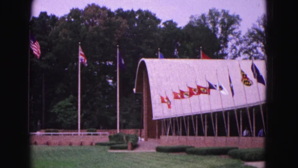 1964 Williamsburg frame showing a curved-roof building lined with flags, with tall flagpoles, trees, lawn, and hedges.