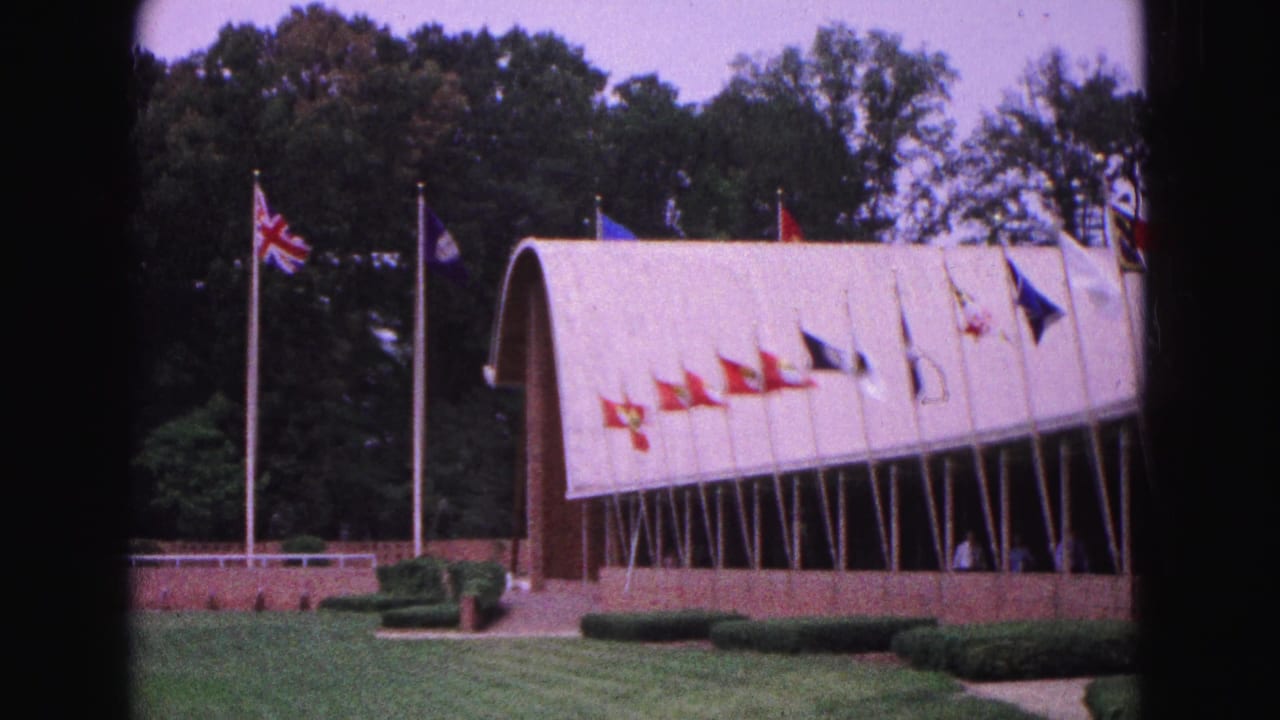 Flags on poles outside a pale arched building with lawn and trees.