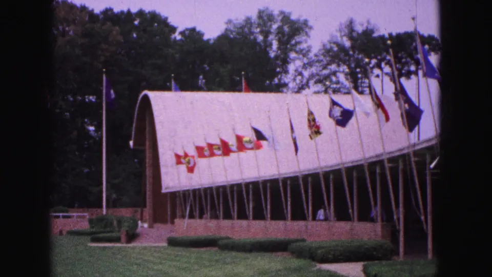 1964 Williamsburg frame with multiple flags along the front of an arched-roof building, with lawn, hedges and trees visible.