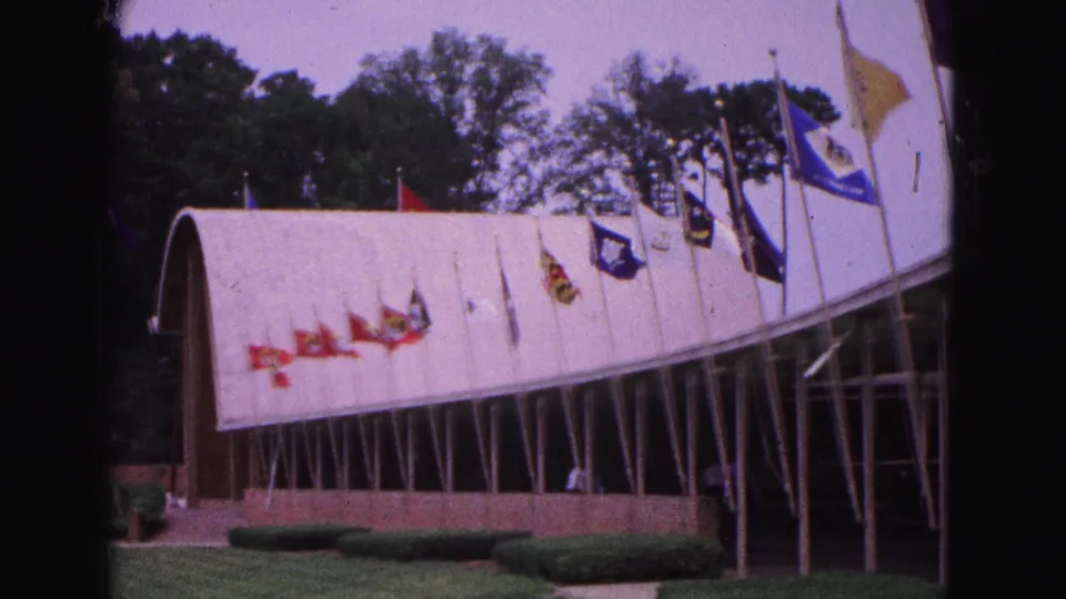 1964 Williamsburg 8mm frame showing a white arched roof, flagpoles, glass windows, hedges and a manicured lawn.