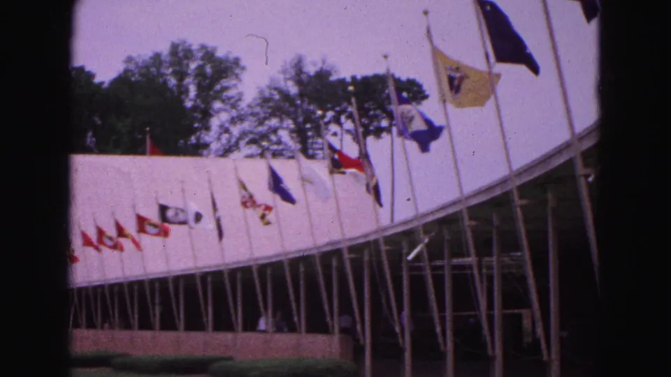 1964 Williamsburg frame with flags blowing near an arched building, glass facade, lawn edge and trees in the background.