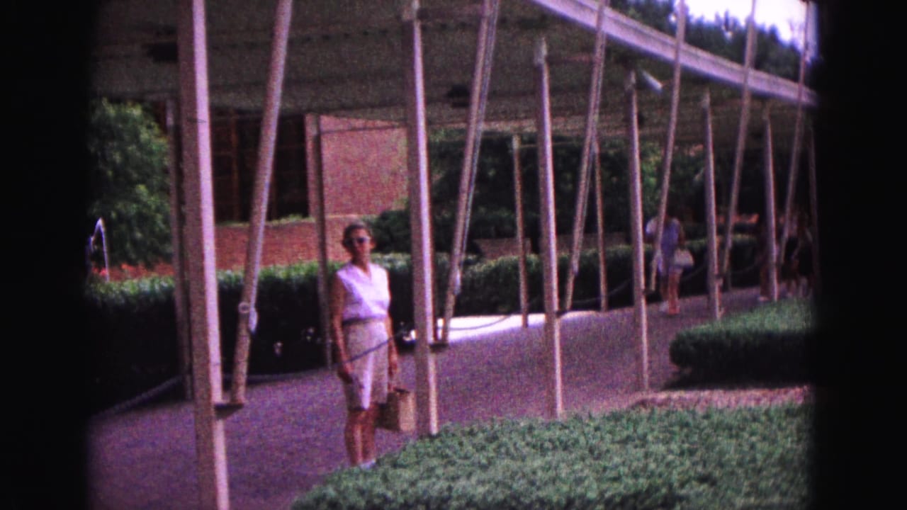 Woman in white stands beside flagpoles near a curved walkway.
