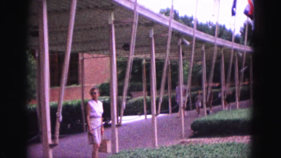 Visitor-shot 8mm frame of a woman in white near the Williamsburg flag display, with slender poles, hedges, walkway, and modernist roofline visible.
