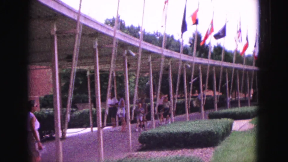 Wide 1964 Williamsburg frame showing visitors on a walkway under the circular flag display, with lawn, hedges, flags, and pavilion supports.