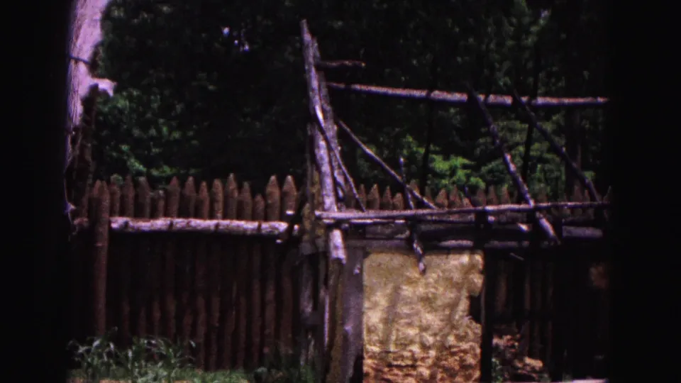 1964 Williamsburg frame showing a wooden fence, rough timber framework, greenery, and part of a rustic wall or small outbuilding.