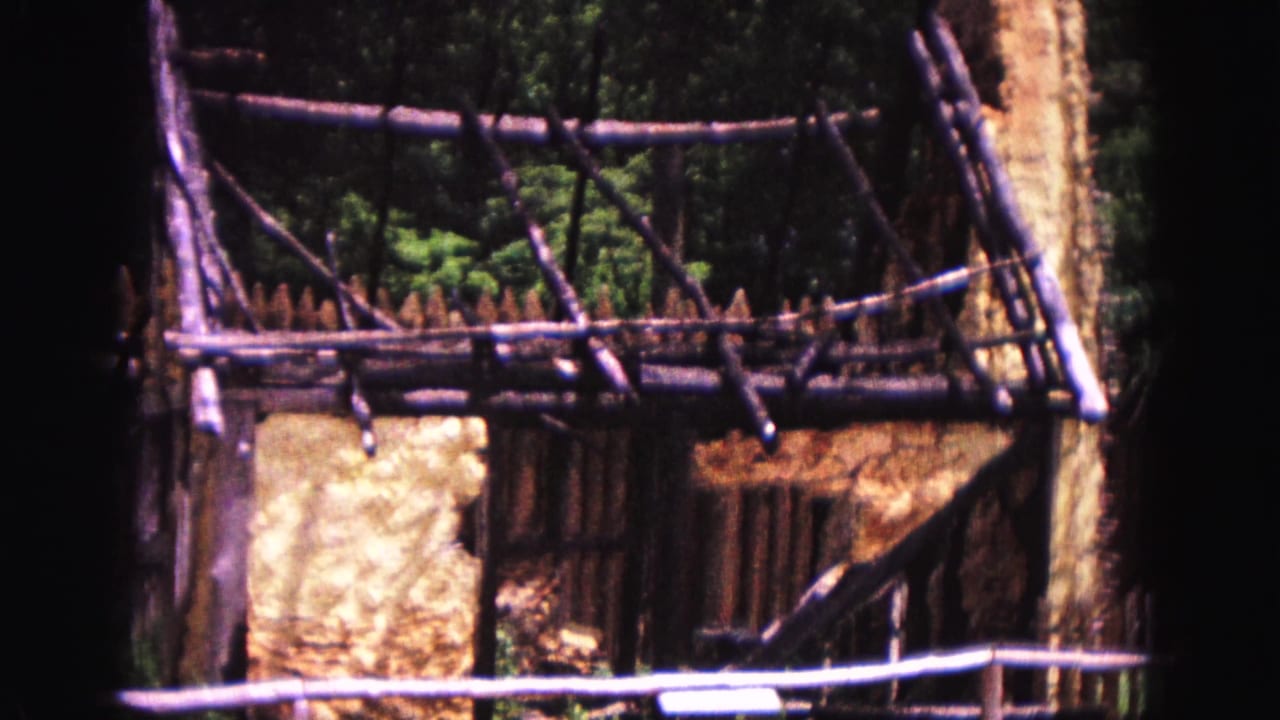 1964 Williamsburg frame with rustic wooden bracing, palisade fencing, and an earth-colored wall in a wooded setting.