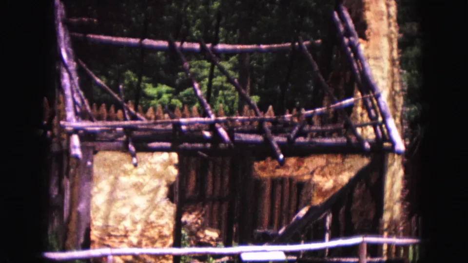1964 Williamsburg frame with rustic wooden bracing, palisade fencing, and an earth-colored wall in a wooded setting.