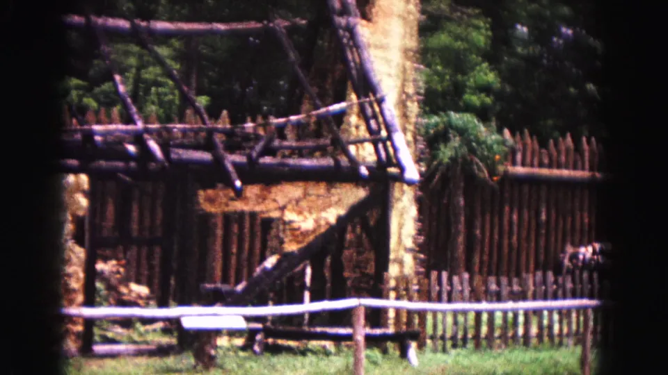 1964 Williamsburg home-movie view of palisade fencing, timber supports, and grass around a reconstructed rustic building.