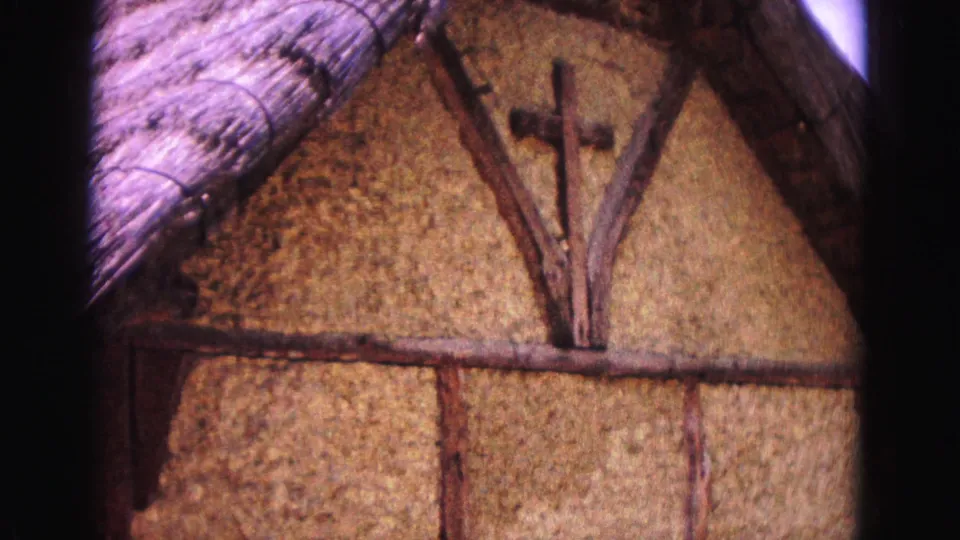 1964 Williamsburg footage showing a thatched-roof gable, rough wall surface, exposed timber framing, and a wooden cross.