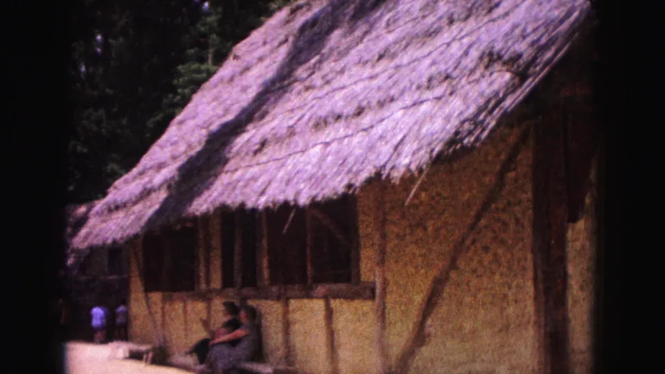 Wider 1964 Williamsburg frame showing the thatched building, timber supports, shaded window area, trees, and seated visitors nearby.