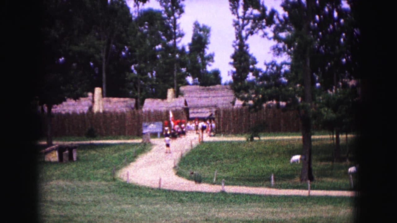 Visitors walk along a curving path toward a fenced historic-site entrance with trees and lawn.