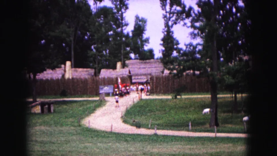 1964 Williamsburg visitor-shot view of a path leading toward a fenced entrance, with tourists, trees, grass, and small white markers visible.