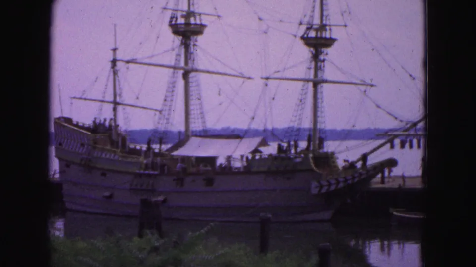 1964 Williamsburg home-movie frame showing the side of a docked wooden sailing vessel, its masts and rigging, pier structures, water, and greenery in front.