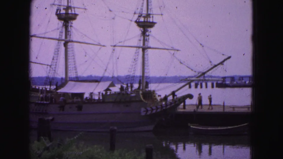 1964 Williamsburg waterfront frame with a rigged wooden vessel, gangway, pier, small tourist figures, calm water, and distant shoreline.