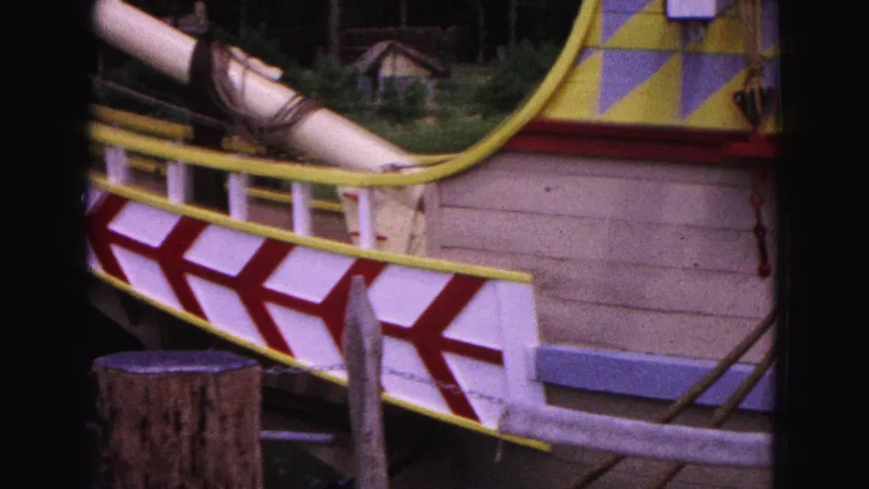 Close 8mm frame of the Williamsburg replica ship side, showing red and white hull pattern, yellow rail and wooden dock posts.