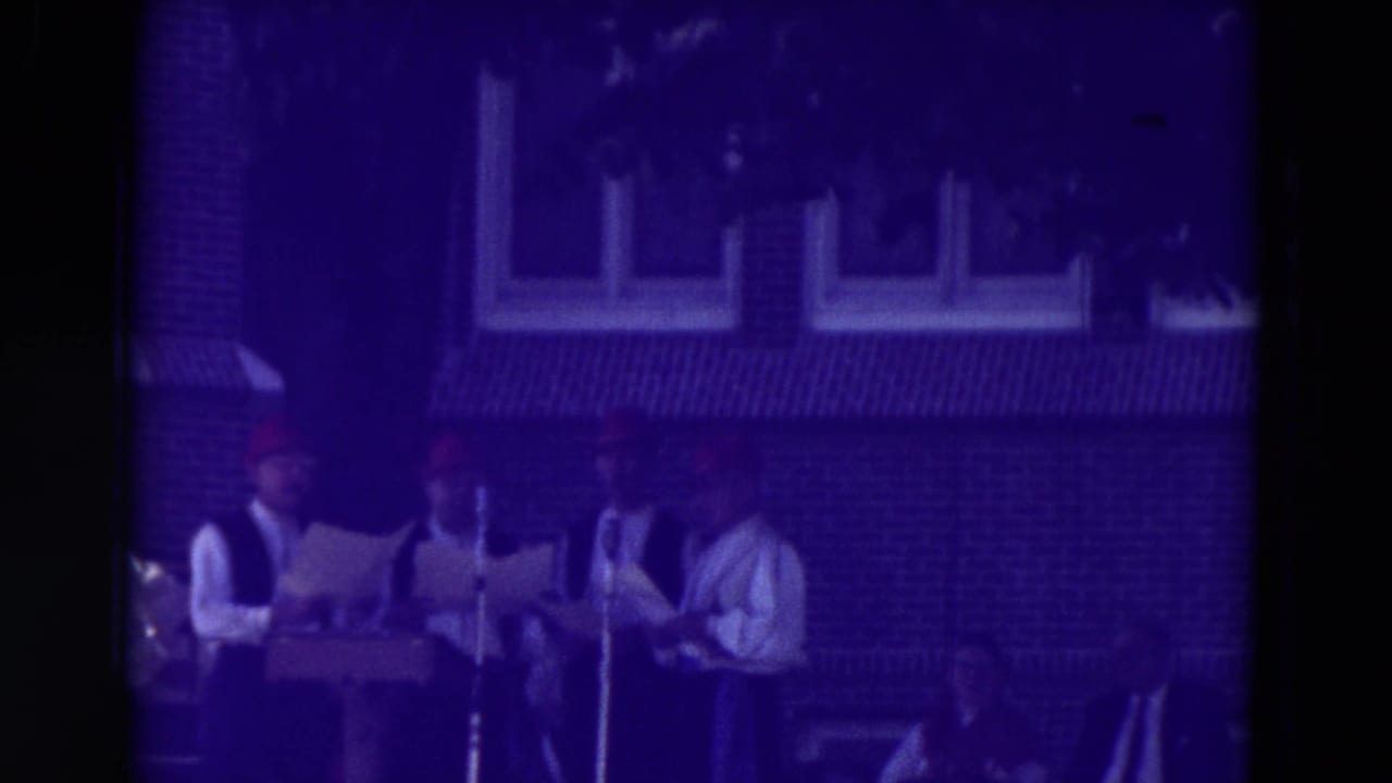 1965 Clarkson, Nebraska home-movie frame showing musicians in white shirts and dark vests at standing microphones before a brick building.