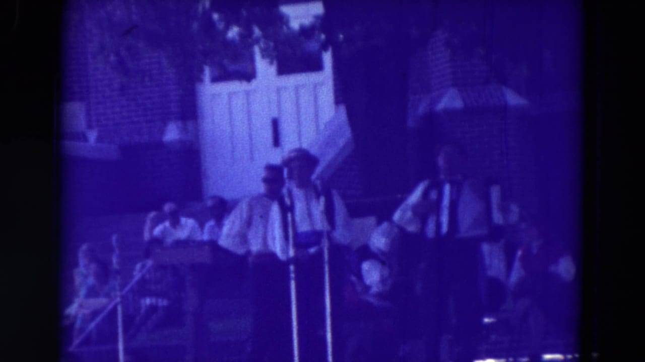 1965 Clarkson, Nebraska frame of outdoor musicians on a small stage, including an accordionist, with spectators and a white doorway behind.