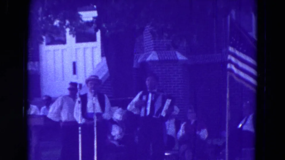 1965 Clarkson, Nebraska home-movie frame showing stage performers, an accordionist, seated audience members, and an American flag at the edge.