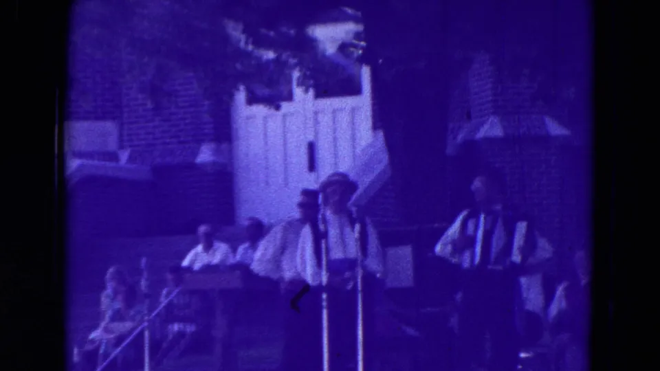 1965 Clarkson, Nebraska frame of two performers at standing microphones, one holding an accordion, in front of spectators and a brick building.