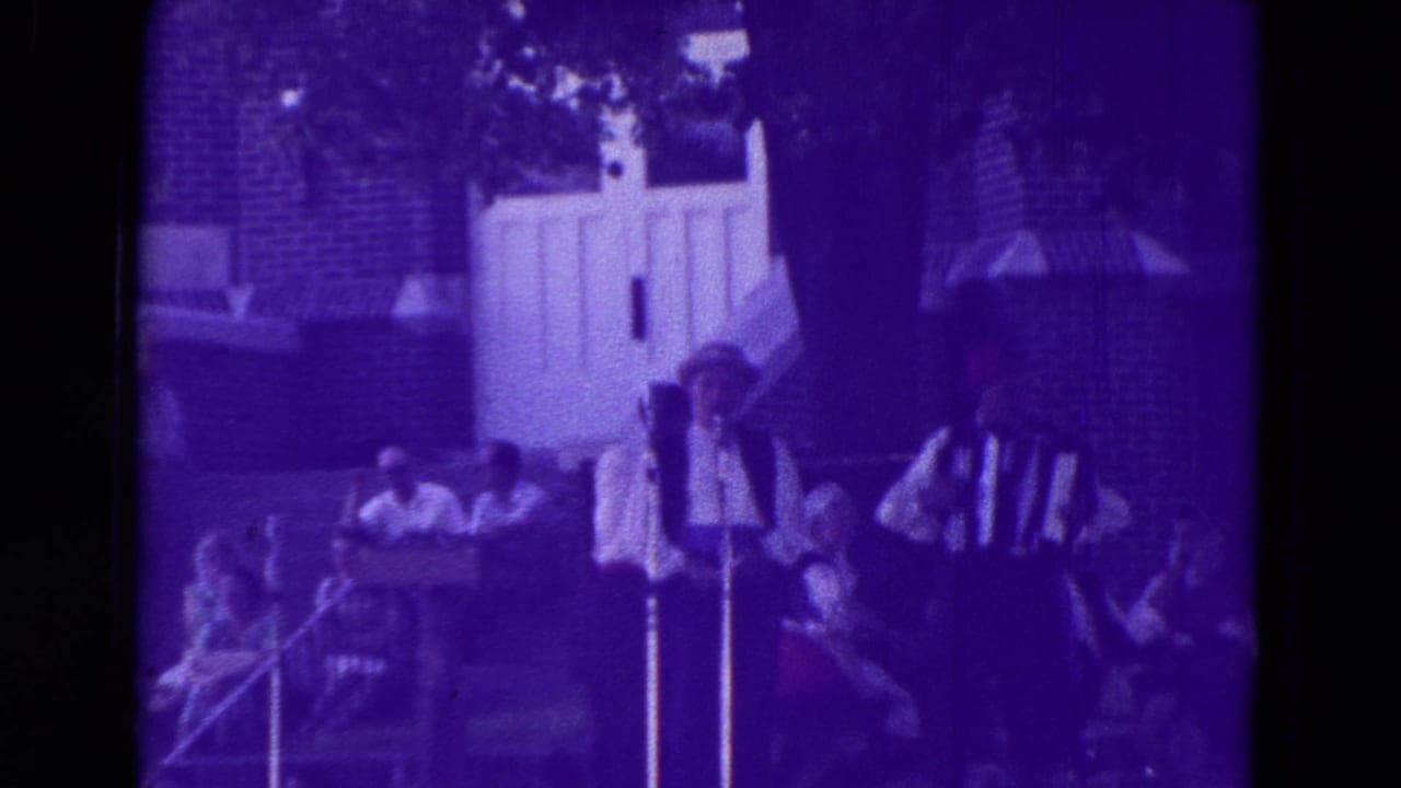 Outdoor street performance in Clarkson, Nebraska, with two figures at microphones and seated onlookers visible through purple-tinted 8mm film.