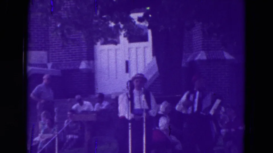 Purple-tinted 8mm frame of an outdoor band performance in Clarkson, Nebraska, with an accordion player and singer near microphones.