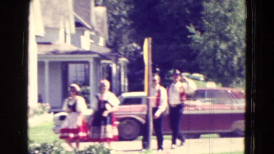 Clearer 1965 Clarkson, Nebraska frame showing people in folk-style clothing near a red car and residential street after the performance scene.