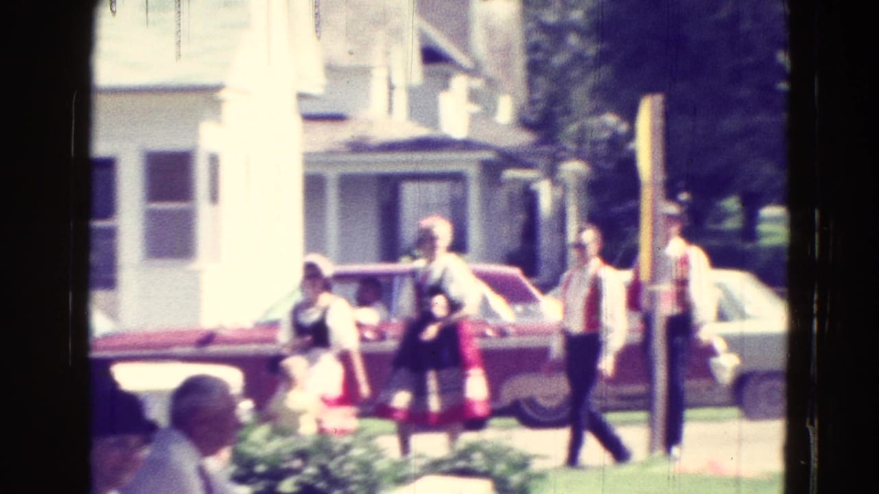 People gather near a red car and white house, with women in white blouses and red skirts.