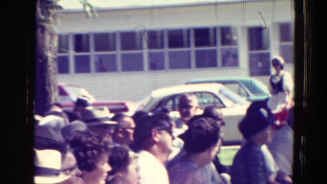 A crowd of spectators sits and stands near parked cars and a low building.
