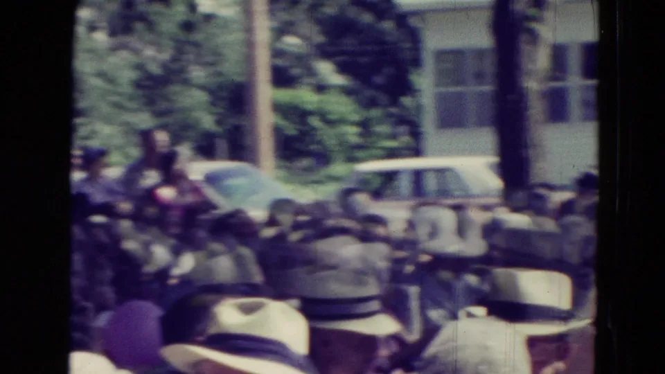 Soft 8mm frame from Clarkson, Nebraska showing a crowd seated outdoors near trees, cars, and a background building in 1965.