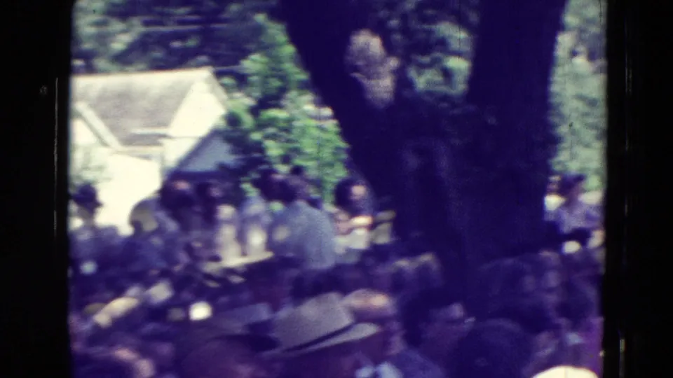 1965 Clarkson, Nebraska home-movie frame showing a shaded outdoor crowd seated beneath trees with houses in the background.