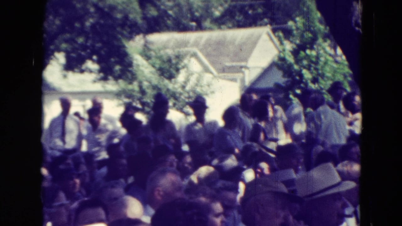 1965 Clarkson, Nebraska home-movie frame showing community members gathered in a shaded park with houses behind them.