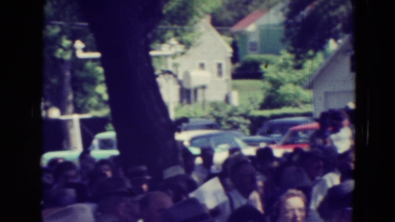 Crowd at an outdoor Clarkson, Nebraska meeting with tree trunks, parked cars, and houses visible beyond the gathering.