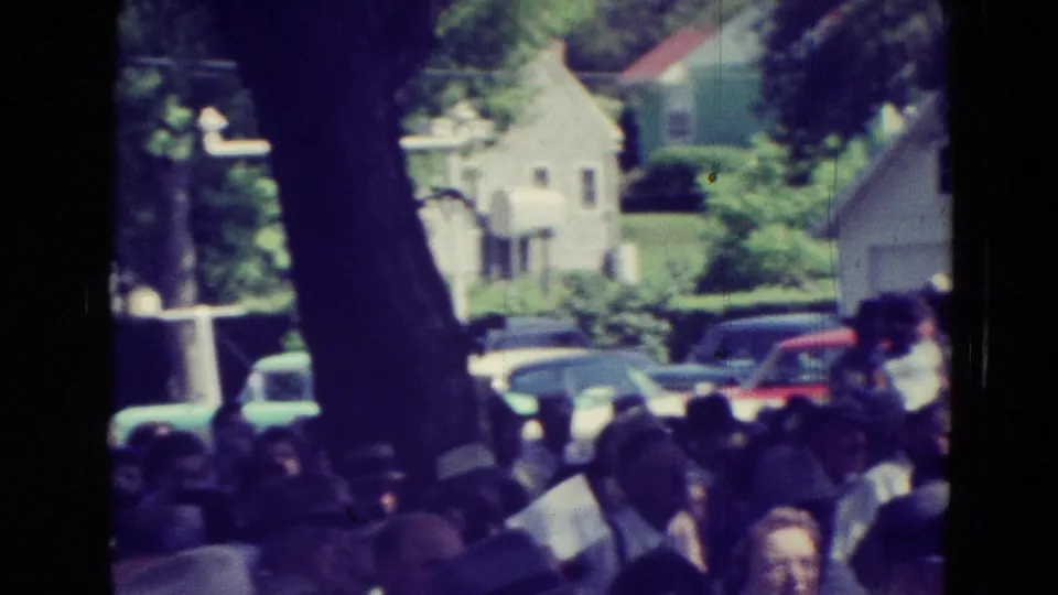 Crowd at an outdoor Clarkson, Nebraska meeting with tree trunks, parked cars, and houses visible beyond the gathering.