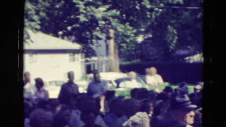 Community members stand near the edge of a 1965 Clarkson, Nebraska park gathering, with cars and houses in the background.