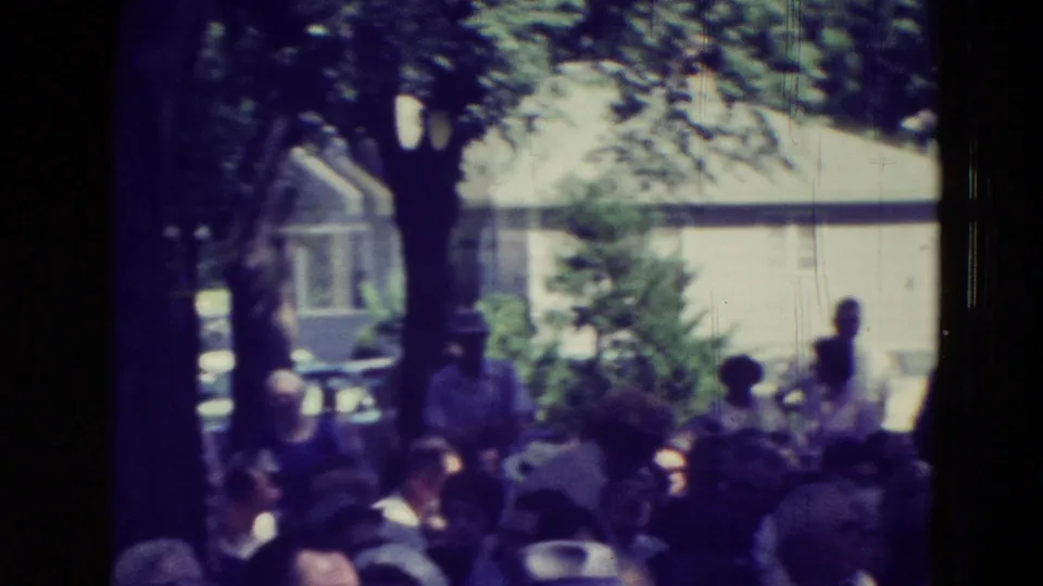 Shaded park crowd in Clarkson, Nebraska, with a large tree, nearby house, and community members facing the meeting area.