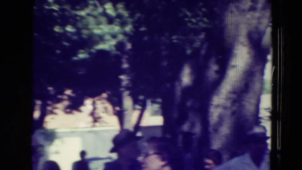 In 1965 Clarkson, Nebraska, adults in summer clothing move through a tree-lined outdoor area in soft, blurred 8mm footage.