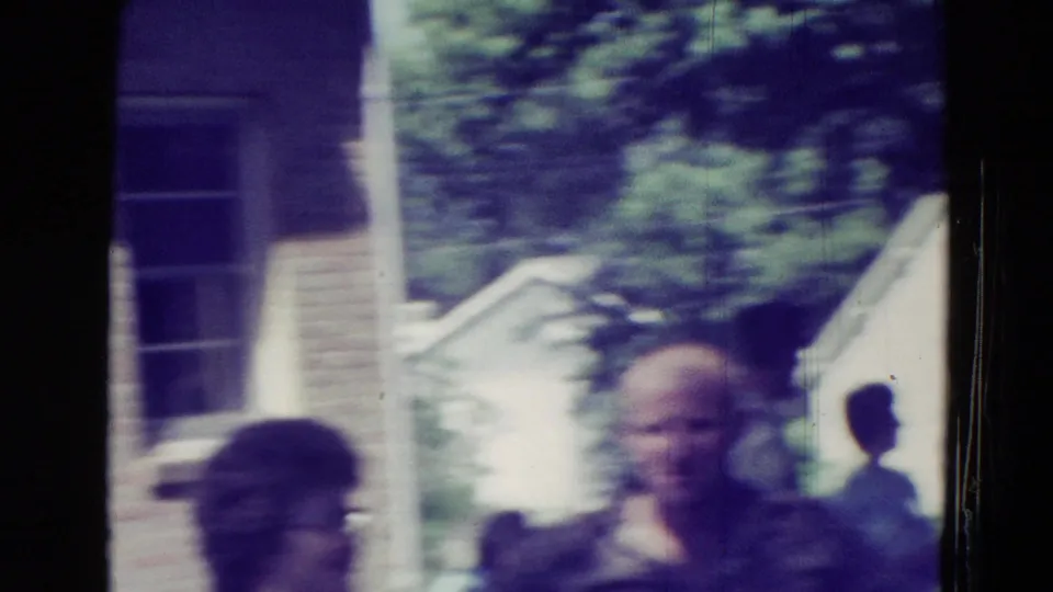 A 1965 Clarkson, Nebraska frame shows adults near a house-like building, porch steps, and trees in handheld 8mm blur.