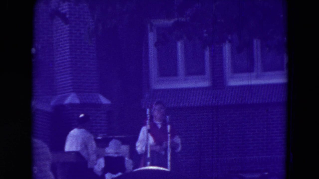 1965 Clarkson, Nebraska home-movie frame showing fair workers and seated visitors near a brick building, heavily tinted purple by aged film color shift.