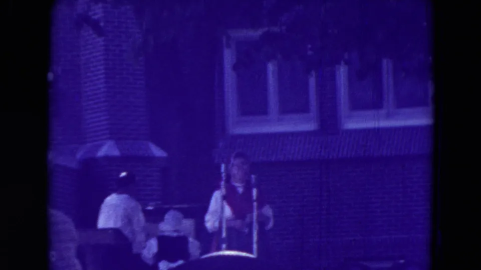 1965 Clarkson, Nebraska home-movie frame showing fair workers and seated visitors near a brick building, heavily tinted purple by aged film color shift.