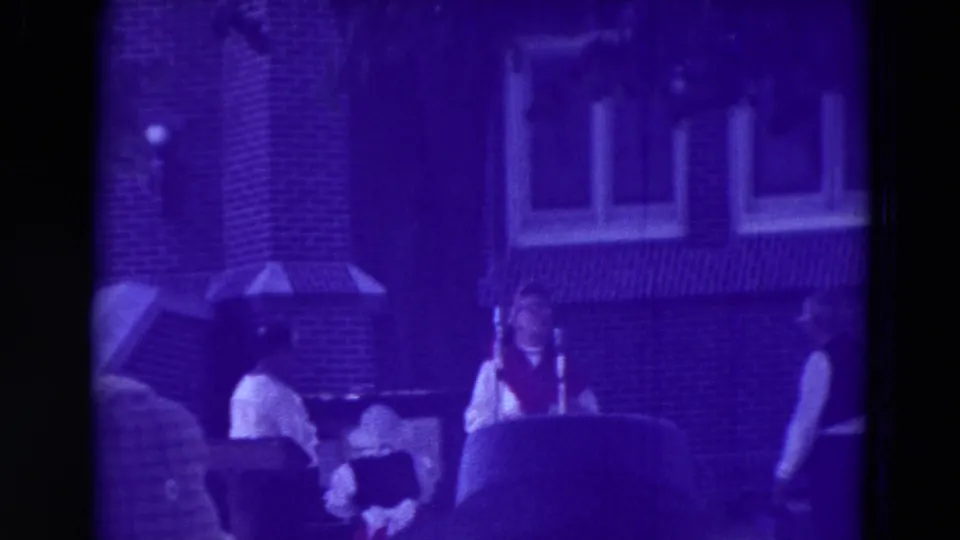 1965 Clarkson, Nebraska carnival footage shows workers in white shirts and vests beside seated visitors near a brick building and canopy.