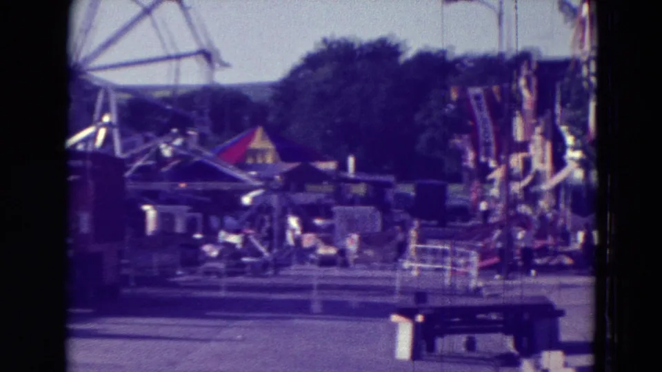 1965 Clarkson, Nebraska carnival frame showing a Ferris wheel structure, tents, benches, trees, and ride equipment under soft 8mm color shift.