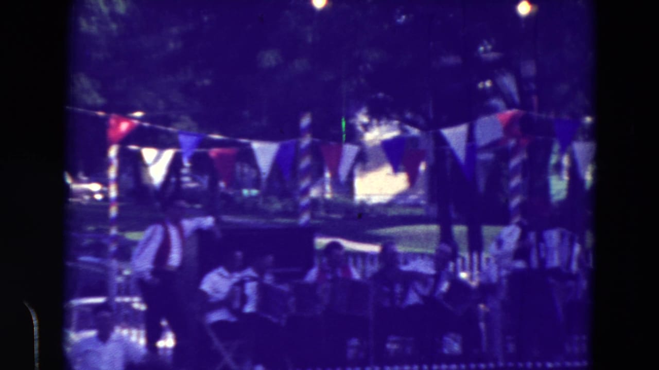 Home-movie view of a decorated Clarkson, Nebraska festival area with accordion players, bunting, and people seated near a fence.