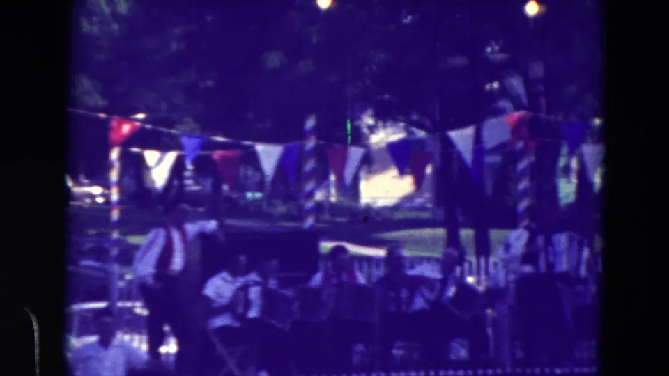Home-movie view of a decorated Clarkson, Nebraska festival area with accordion players, bunting, and people seated near a fence.
